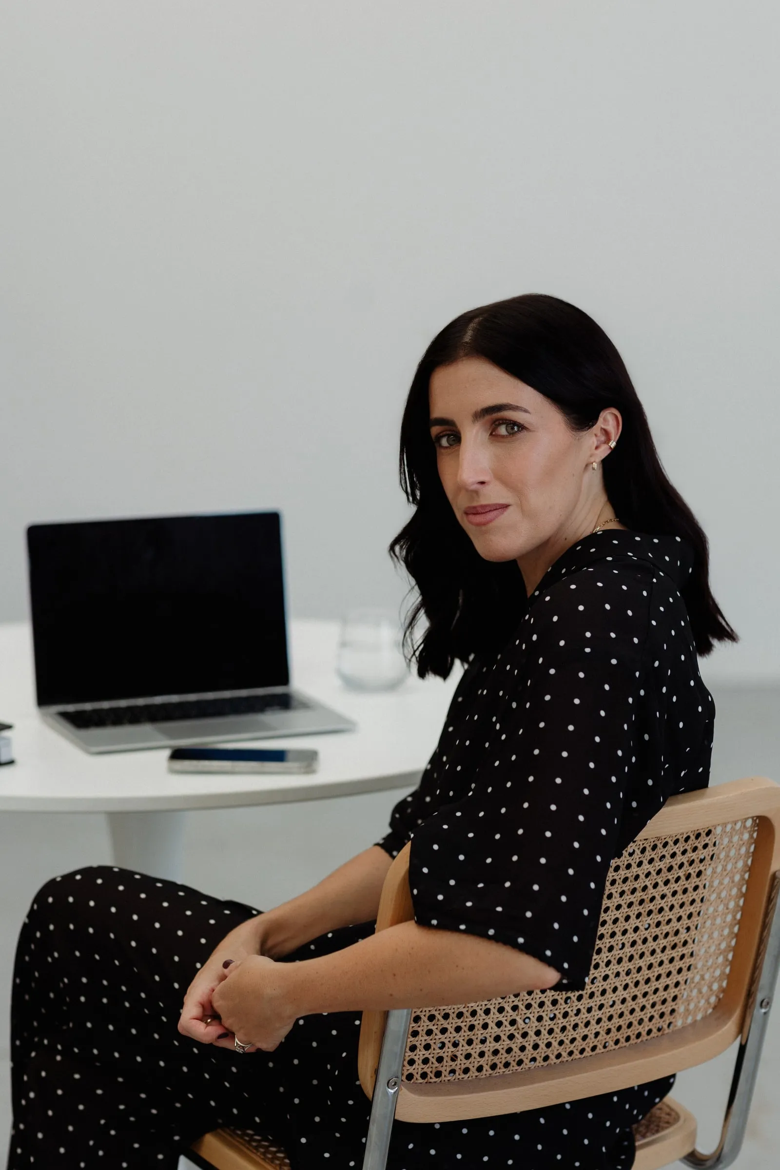 Liana McLeod looking over her shoulder from a cane chair, beside a table with her laptop and phone.