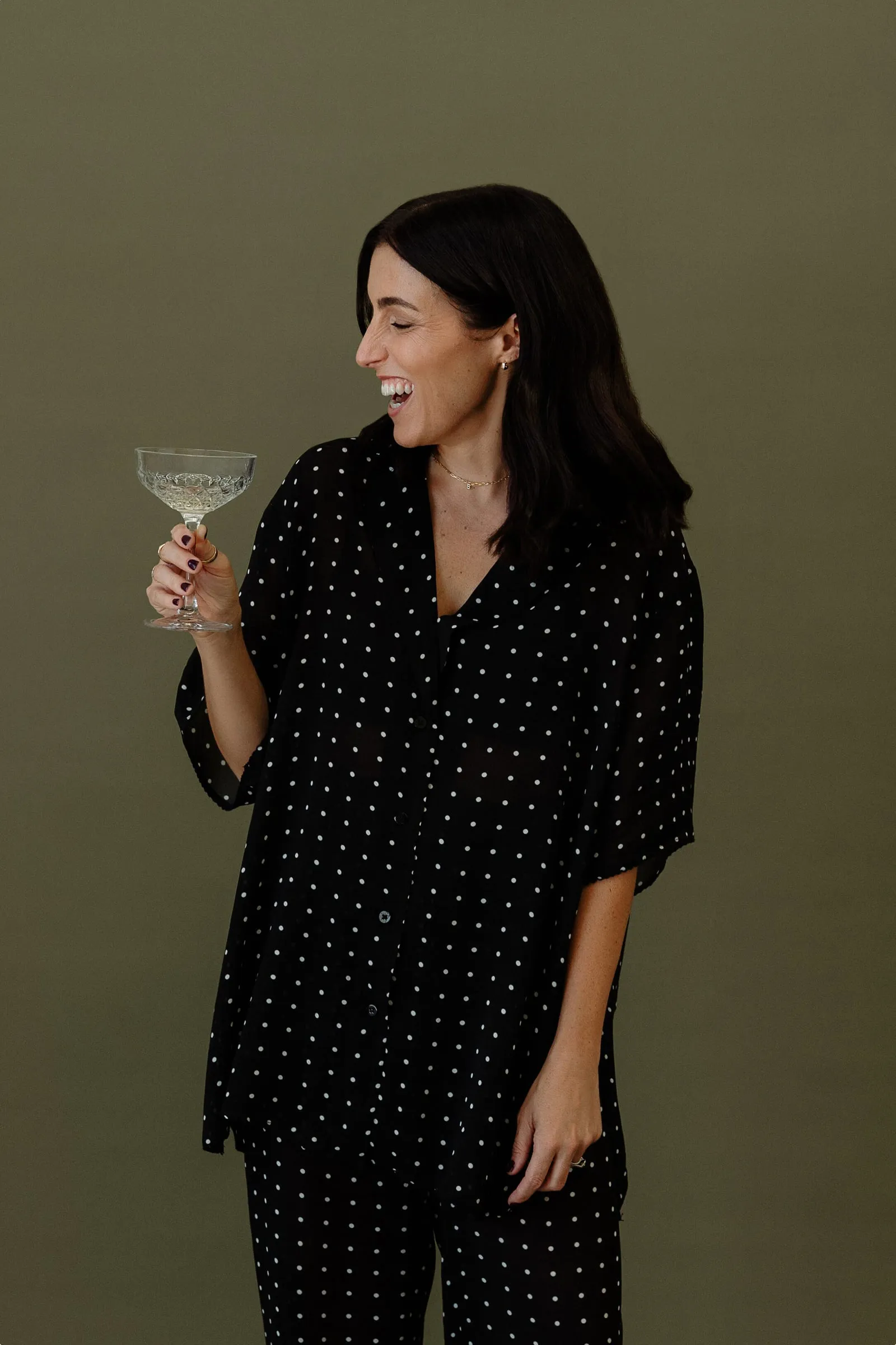 Liana McLeod laughing, holding a coupe glass, against an olive backdrop.