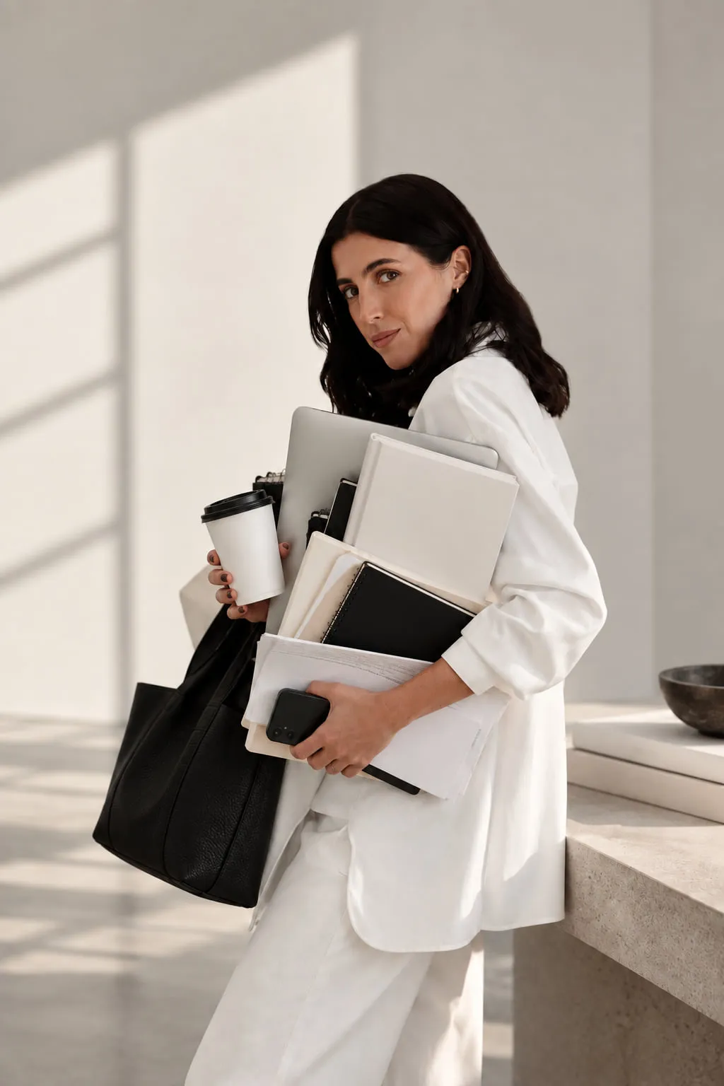 Liana McLeod leaning against a stone bench, holding a coffee, a laptop and an armful of notebooks and folders.