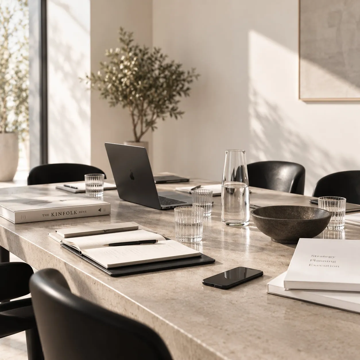 A sunlit meeting table set with notebooks, a laptop, glasses and a carafe of water.
